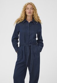 Frau mit lockigem blondem Haar, die einen dunkelblauen Jeans-Overall mit Kragen, Frontknöpfen, gebundenem Gürtel und Händen in den Taschen trägt.