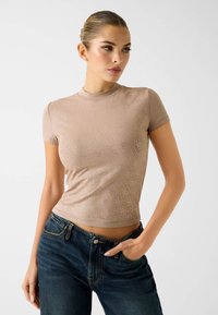 Dopasowany beżowy top z krótkim rękawem o fakturowanym wzorze diamentu; w zestawieniu z ciemnoniebieskimi jeansami i ręką w kieszeni, podkreślającą wzór topu.