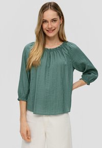 Groene, lichtgewicht blouse met een textuurafwerking, gegolfde halslijn en driekwart mouwen. Gepaard met lichtgekleurde broeken.
