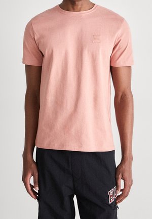 Man draagt een zalmroze T-shirt met korte mouwen en een klein logo op de borst, en zwarte broek met een wit-rode "HUGO" badge die is vastgenaaid.