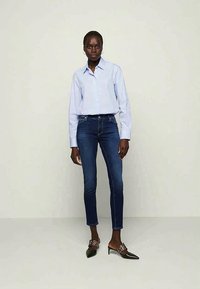 Camisa de botão azul clara, jeans skinny azul escuro, mules pretas com detalhes decorativos, de pé contra um fundo neutro.