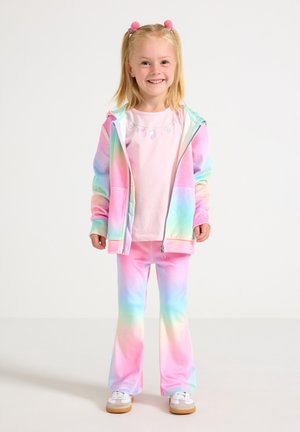 Jeune fille souriante debout, portant un sweat à capuche et un pantalon tie-dye arc-en-ciel pastel, un t-shirt rose clair, des baskets blanches, avec des pompons roses dans les cheveux.