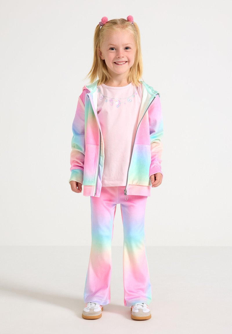 Jeune fille souriante debout, portant un sweat à capuche et un pantalon tie-dye arc-en-ciel pastel, un t-shirt rose clair, des baskets blanches, avec des pompons roses dans les cheveux.
