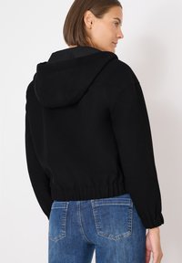 Sudadera corta negra con capucha ajustable, dobladillo acanalado y mangas relajadas. Combinada con jeans de mezclilla azul, mostrando un estilo casual.