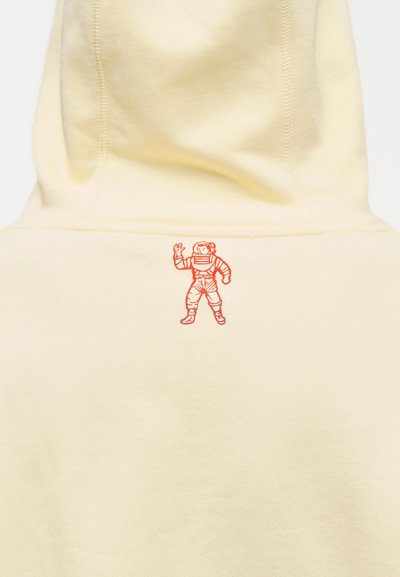 Κίτρινο φούτερ με κουκούλα που διαθέτει κόκκινο κεντημένο αστροναύτη στην πλάτη, κατασκευασμένο από απαλό ύφασμα με ορατές ραφές.