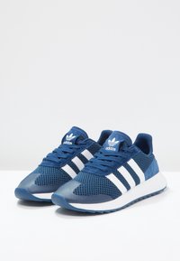 Námořnické modré sportovní boty s síťovinovým svrškem, bílými pruhy, polstrovaným límcem a texturovanou gumovou podrážkou. Disponují aerodynamickým designem a polstrovaným interiérem.