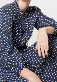 Robe noire avec un motif géométrique bleu et blanc, encolure froncée et manches ballon, avec des poignets élastiques et un détail à cordon.