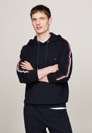 Sudadera azul marino con acentos en rayas rojas y blancas en las mangas, con un bolsillo frontal y capucha con cordón. Textura de tela suave.