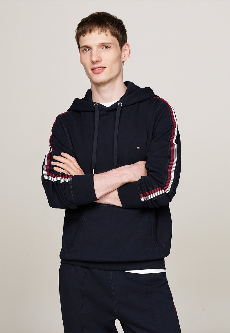 Sudadera azul marino con acentos en rayas rojas y blancas en las mangas, con un bolsillo frontal y capucha con cordón. Textura de tela suave.