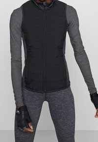 Svart ärmlös puffer väst med horisontell sömnad och dragkedja fram, lager på lager över en grå långärmad topp och texturerade grå leggings.