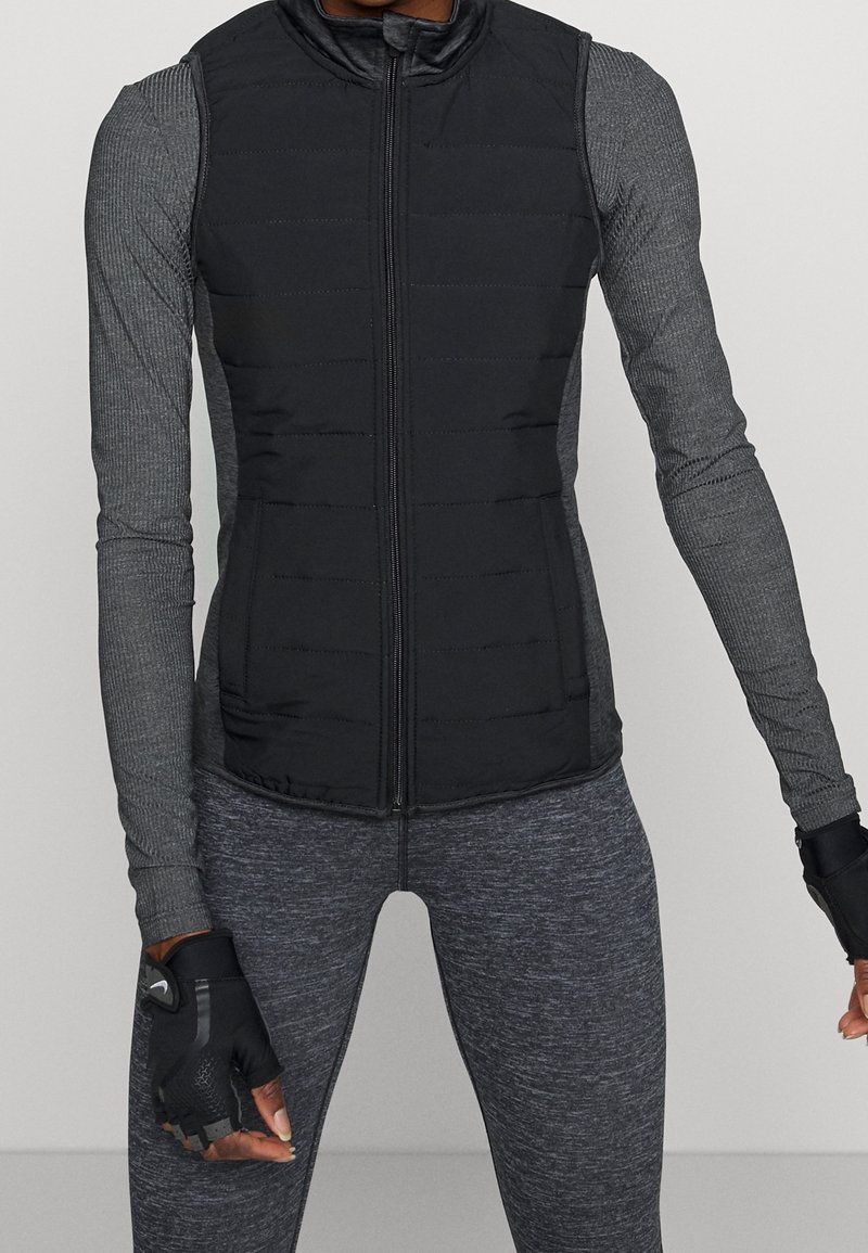 Svart ärmlös puffer väst med horisontell sömnad och dragkedja fram, lager på lager över en grå långärmad topp och texturerade grå leggings.