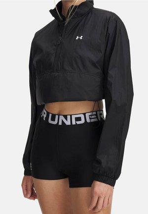 Person iført en sort afkortet lynlåstrøje og sort shorts med højtaljet bred elastisk taljebånd med mærke.