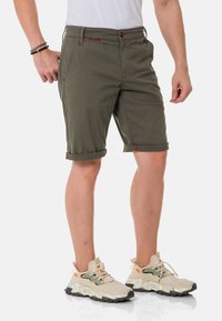 Olijfgroene shorts met omgeslagen manchetten, voorzien van contrasterende rode stiksels en een ritssluiting. G gecombineerd met beige gestructureerde sneakers en een wit shirt.