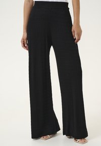Pantalons noirs à jambes larges avec une texture côtelée et une taille élastique. Le tissu est doux, et les pantalons descendent jusqu'aux chevilles, associés à des chaussures légères.