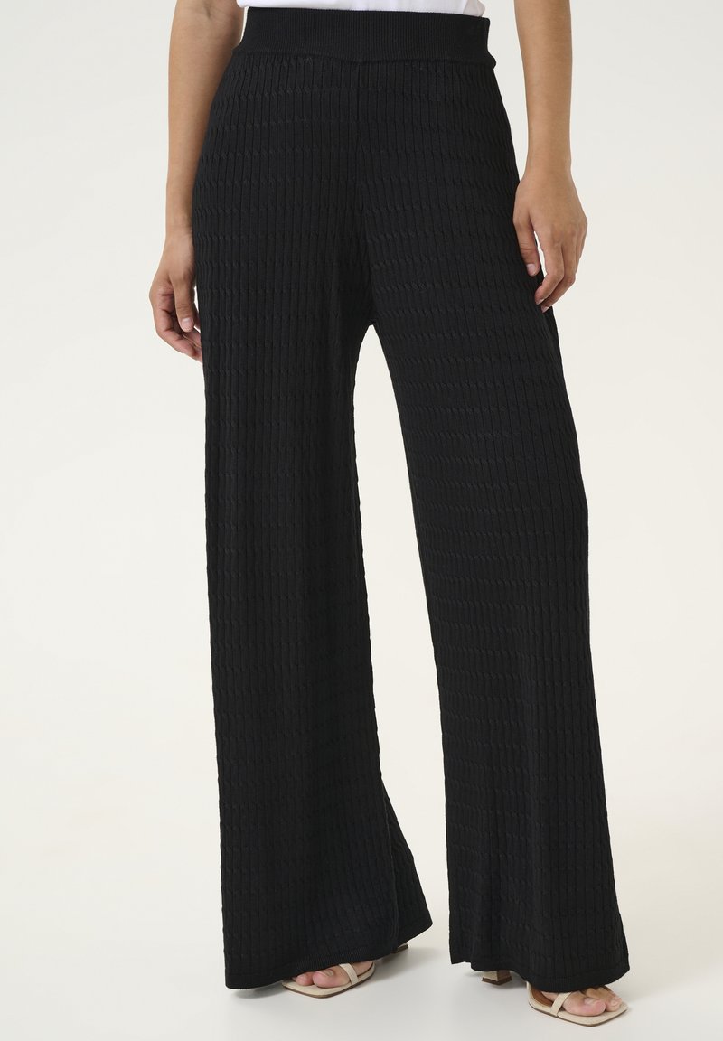 Pantalons noirs à jambes larges avec une texture côtelée et une taille élastique. Le tissu est doux, et les pantalons descendent jusqu'aux chevilles, associés à des chaussures légères.