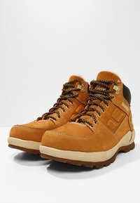 Bottes de randonnée en cuir tan avec un col rembourré, des decorations dorées, des lacets noirs et jaunes, et une semelle extérieure contrastante blanche et brune.