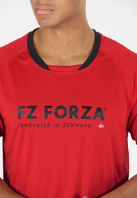 Punainen urheilu paita lyhyillä hihoilla, musta kaula-aukko, jossa on suuri musta logo "FZ FORZA" ja teksti "INNOVATED IN DENMARK."