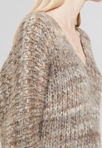 Kvinna som bär en grovstickad tröja i blandade beige, bruna och vita nyanser med V-ringning och ett fint guldhalsband.