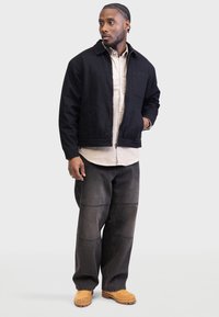Sort zip-up jakke med høj krave og frontlomme, parret med løse mørkegrå jeans og brune fodtøjer, sat mod en ensfarvet baggrund.