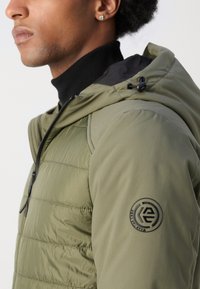 Doudoune de couleur olive, avec une fermeture éclair à l'avant, une capuche réglable et un logo noir rond sur l'épaule gauche. Texture lisse.