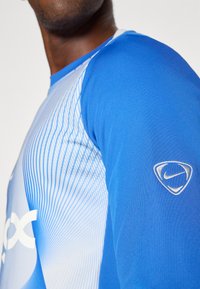 Hosszú ujjú sportpóló kék és fehér színben, ami átmenetes mintázattal és átlós vonalakkal rendelkezik, valamint egy fehér Nike logóval az ujjon.