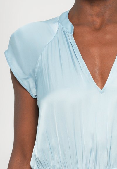 Lichtblauw, zijden top met een V-hals en korte, cap sleeves. De stof heeft een zachte glans en heeft subtiele plooien bij de taille.