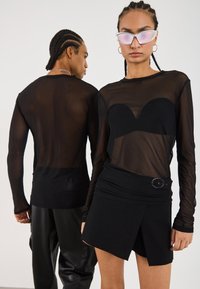 Schwarzes Mesh-Oberteil mit langen Ärmeln und einer schwarzen trägerlosen Innenschicht, kombiniert mit einem enganliegenden schwarzen Miniröckchen mit seitlichem Akzent.