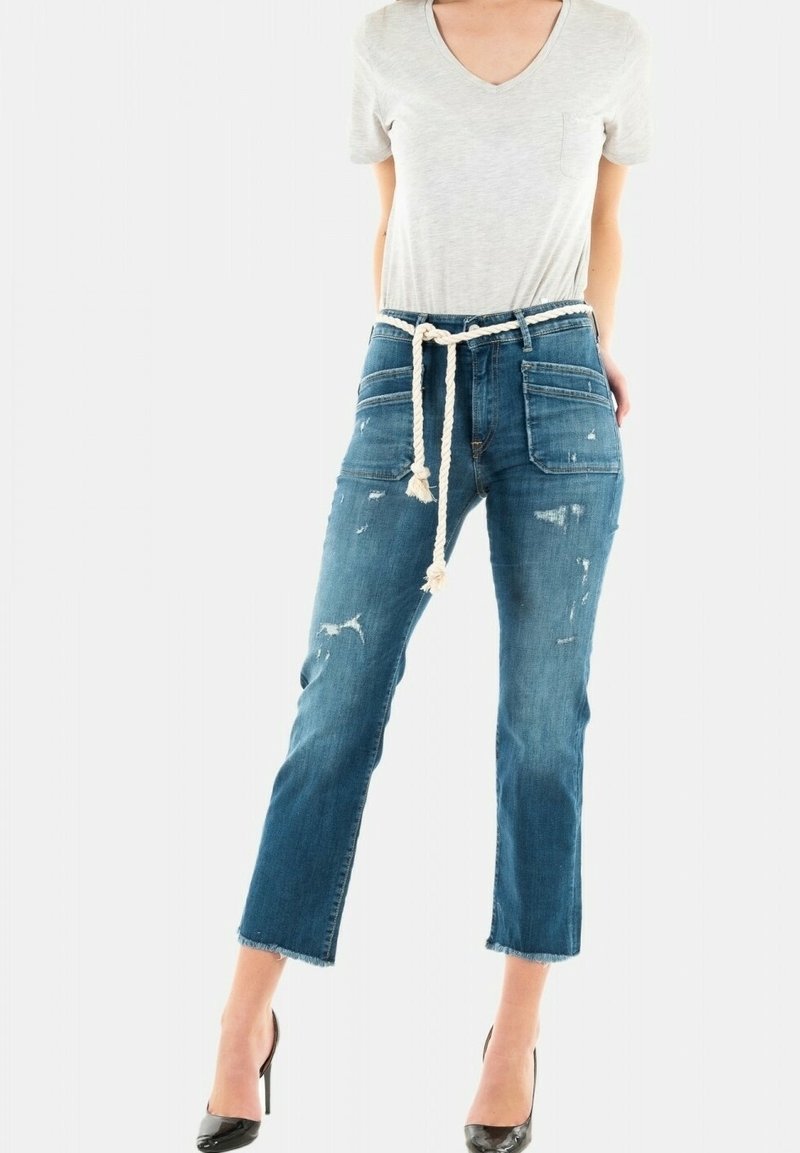 T-shirt gris clair à col en V associé à un jean en denim bleu délavé à taille moyenne, usé, avec une cordelette tressée et une coupe raccourcie. Escarpins noirs à talons aiguilles.