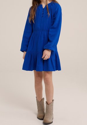 Fille portant une robe bleu à manches longues avec des boutons et des bottines en daim beige, debout devant un fond beige uni.