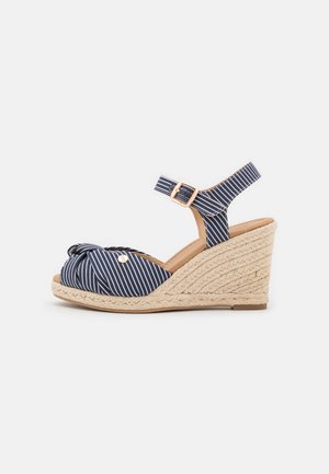 Sandale compensée à rayures bleu marine avec bride à la cheville, boucle dorée, détail noué à l'avant et talon en corde tressée.