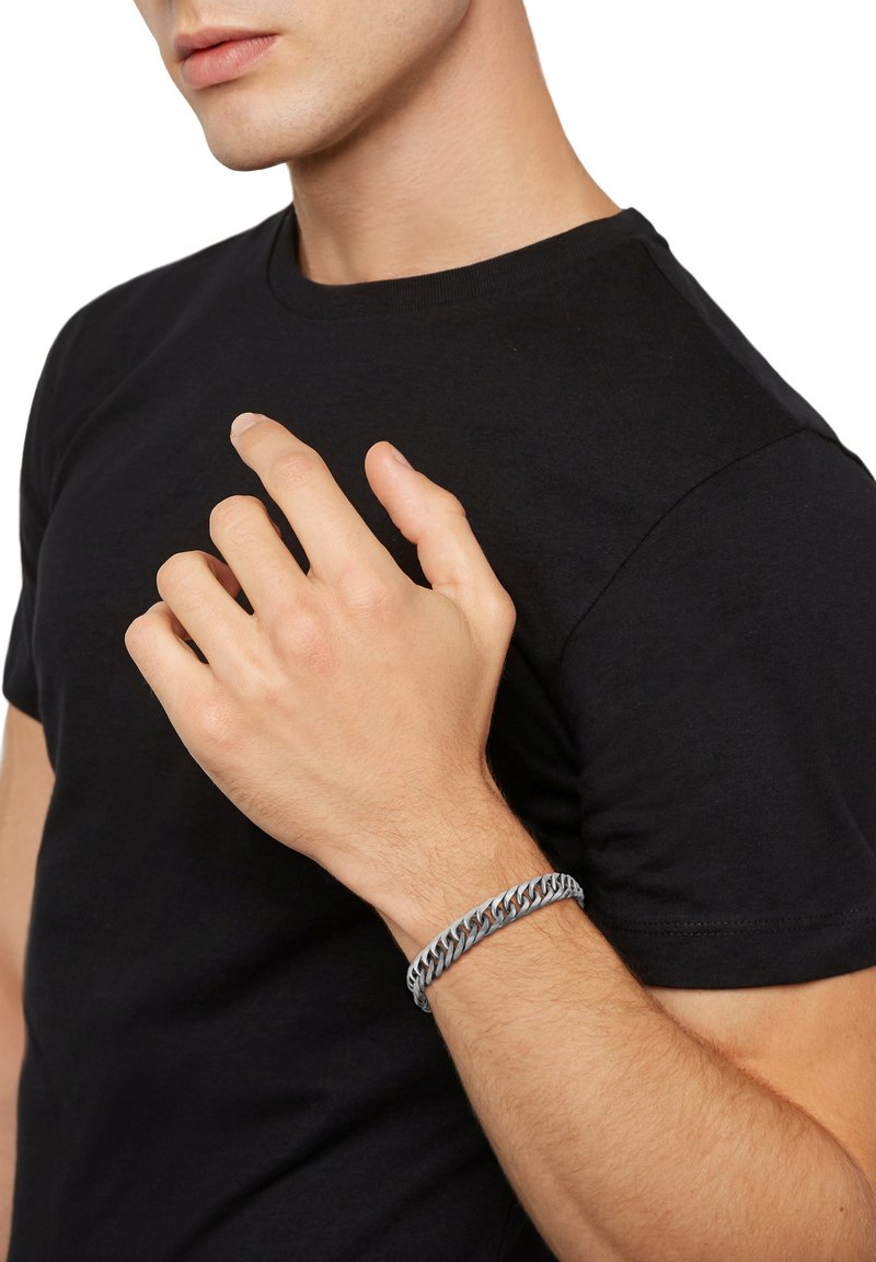 Bracciale in argento su un polso, con maglie intrecciate. Indossato da una persona in t-shirt nera, che mette in risalto la texture e il design del bracciale.