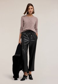 Cardigan tricotat roz deschis cu nasturi, asociat cu pantaloni negri din piele ecologică lucioasă, ținând o haină texturată neagră și purtând mule negre cu toc.