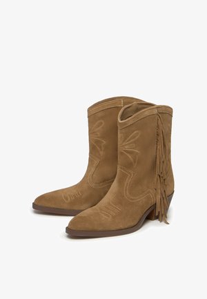 Botines de ante marrón con detalle de flecos y costuras bordadas. Punta afilada, tacón bajo apilado y recortes decorativos en los laterales.
