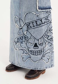 Maxi nederdel i denim med slidte kanter, sort broderet kranie-design med hjerte og tekst. Brune sko med gummisåler er synlige.