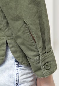 Camisa de manga larga color verde oliva con puños de botón, que presenta un detalle de abertura lateral y una tela texturizada. Vista en primer plano del área de la manga.