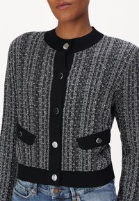 Svart och vit texturerad cardigan med en ribbad krage, silverfärgad knappknäppning och två svarta fickor. Tyget har ett vertikalt randmönster.