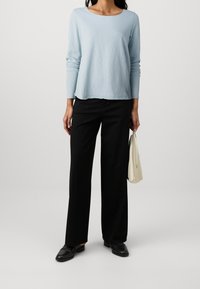 Ljusblå långärmad topp med rund hals, matchad med svarta vida byxor och svarta slip-on skor, hållande en krämfärgad handväska.