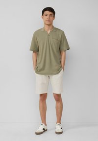 Polo a maniche corte verde oliva con scollatura a V e taschino sul petto, abbinato a pantaloni corti panna e scarpe da ginnastica bianche con dettagli marroni.