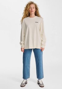 Camiseta de manga larga beige con logo en el pecho, combinada con jeans azul de corte recto y zapatillas marrones/blancas, posando contra un fondo claro.