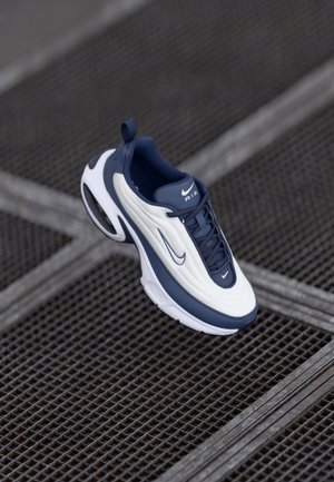 Biało-granatowy but do biegania Nike Air pokazany nad metalową siatką, podkreślający podeszwę i boczne detale.