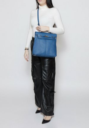 Femme portant un col roulé blanc et un pantalon en cuir noir, tenant un sac bandoulière en cuir bleu avec une fermeture argentée.