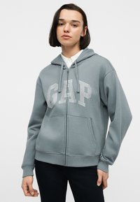 Jeune personne aux cheveux bruns raides portant un sweat à capuche gris GAP zippé sur un t-shirt blanc et un pantalon noir, debout devant un fond uni.
