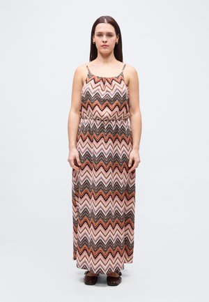 Kvinne står vendt mot kameraet iført en ermeløs maxi-kjole med sikksakkmønster og spaghetti-stropper, samt brune sko, foran en ensfarget hvit bakgrunn.