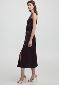 Dunkelbraunes, ärmelloses Kleid mit einem seitlichen Schlitz, goldenen Knopfdetails und einem taillierten Oberteil, das in einen ausgestellt Rock übergeht.