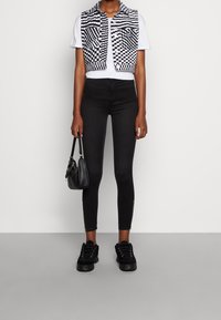 Persona vestida con un chaleco a cuadros blanco y negro sobre una camiseta blanca, jeans ajustados negros con cremalleras, zapatillas negras, sosteniendo un pequeño bolso negro.