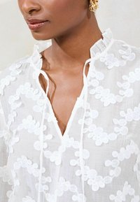 Weißes besticktes Oberteil mit floralen Mustern, einem gerafften Kragen und Krawattendetails am Halsauschnitt. Leichter, transparenter Stoff.