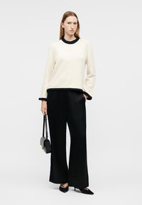 Femme debout portant un pull blanc avec des bordures noires, un pantalon ample noir, des talons noirs, et tenant un petit sac à main noir.