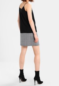 Femme portant un haut noir sans manches avec un dos en dentelle, une mini-jupe à carreaux et des bottines à talons noirs, debout de dos.