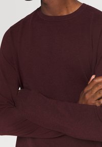 Man med korsade armar som bär en långärmad burgundy stickad tröja mot en enfärgad ljus bakgrund.