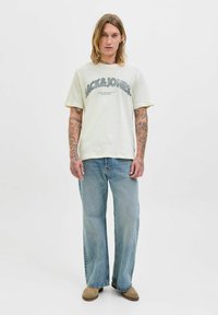 Kremo spalvos medvilninis marškinėliai su "Jack & Jones" grafika. Dėvimi su šviesiai mėlynomis plačiomis kelnėmis ir beigiškomis bateliais, matomos tatuiruotės ant rankų.